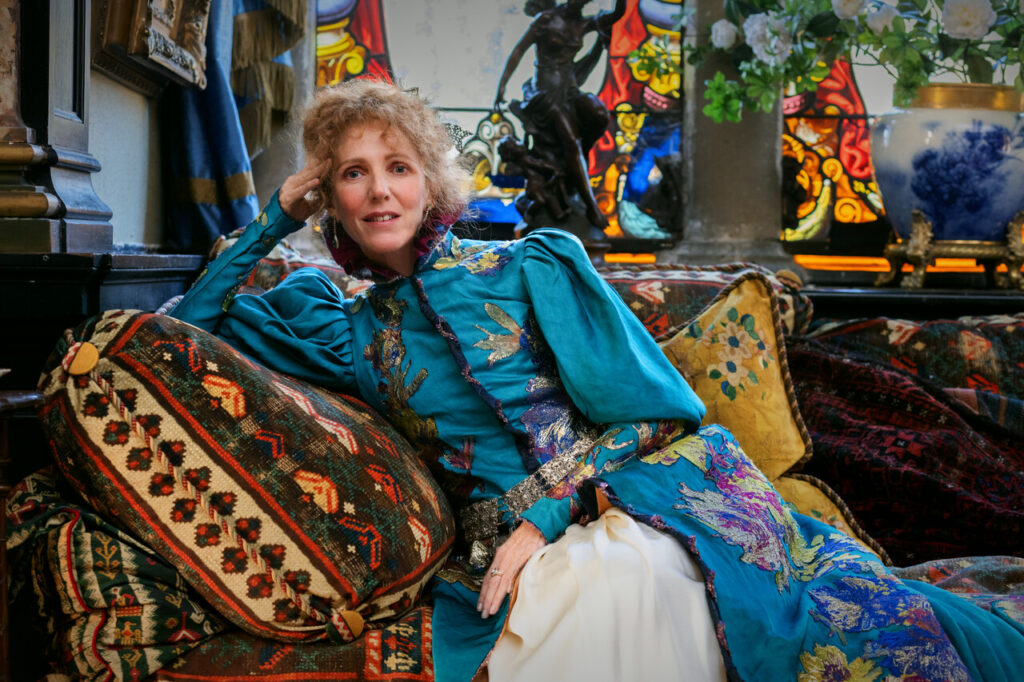 Sandrine Kiberlain como Sarah Bernhardt sentada em sofá, cercada por vitrais e objetos decorativos
