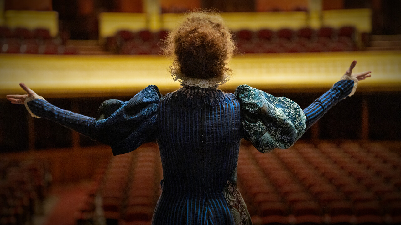 Atriz como Sarah Bernhardt de braços abertos diante da plateia vazia de um teatro
